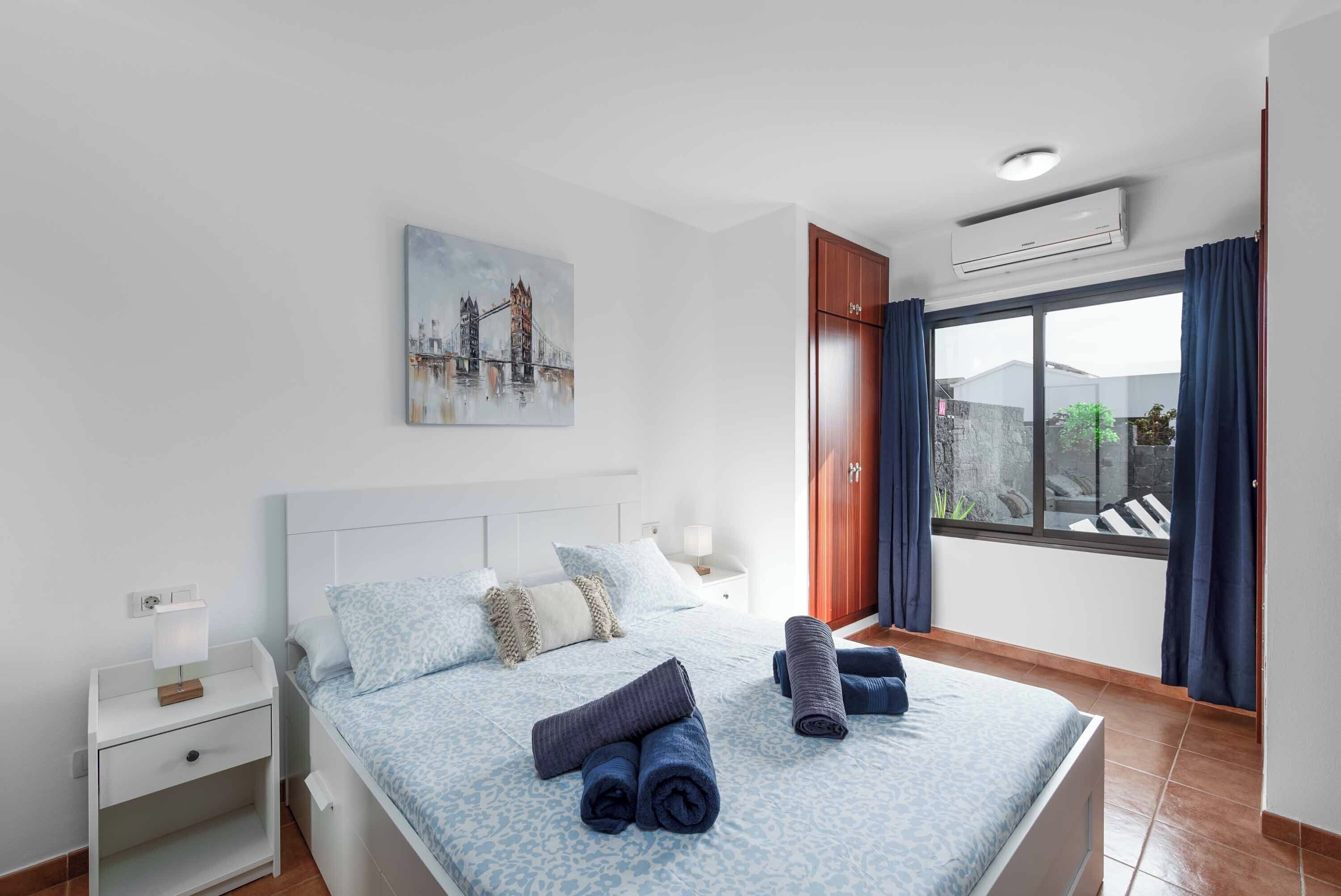 A bedroom with a bed, a nightstand, a painting of a bridge, and a window with curtains.