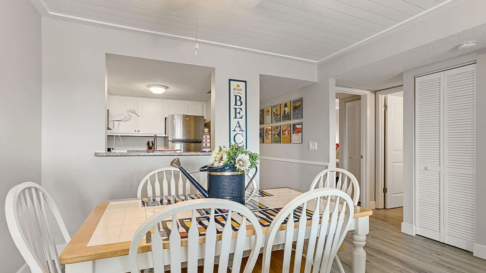 A dining area with a wooden table, white chairs, and a vase with flowers. A kitchen is visible in the background.