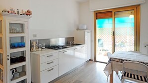 Eine moderne Küche mit weißen Schränken, einem Gasherd und einem Spülbecken. Ein Esstisch mit einem Stuhl und ein Fenster mit Blick ins Freie sind ebenfalls vorhanden.