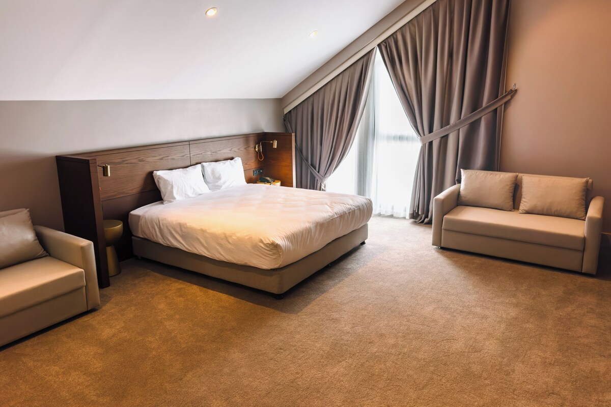 Une chambre d’hôtel moderne avec un grand lit, deux fauteuils, une table de chevet et une fenêtre avec des rideaux.