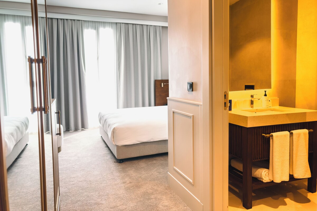 Une chambre d’hôtel avec un grand lit, une table de chevet, une salle de bain avec un lavabo et un miroir, et une vue sur l’extérieur par la fenêtre.