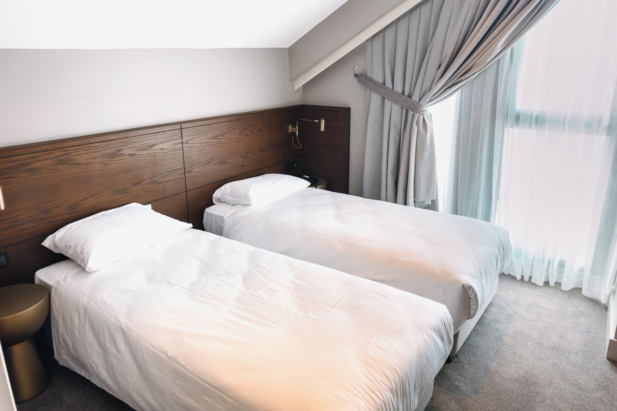 Une chambre d’hôtel avec deux lits simples, une tête de lit en bois, une table de chevet et une grande fenêtre avec des rideaux.