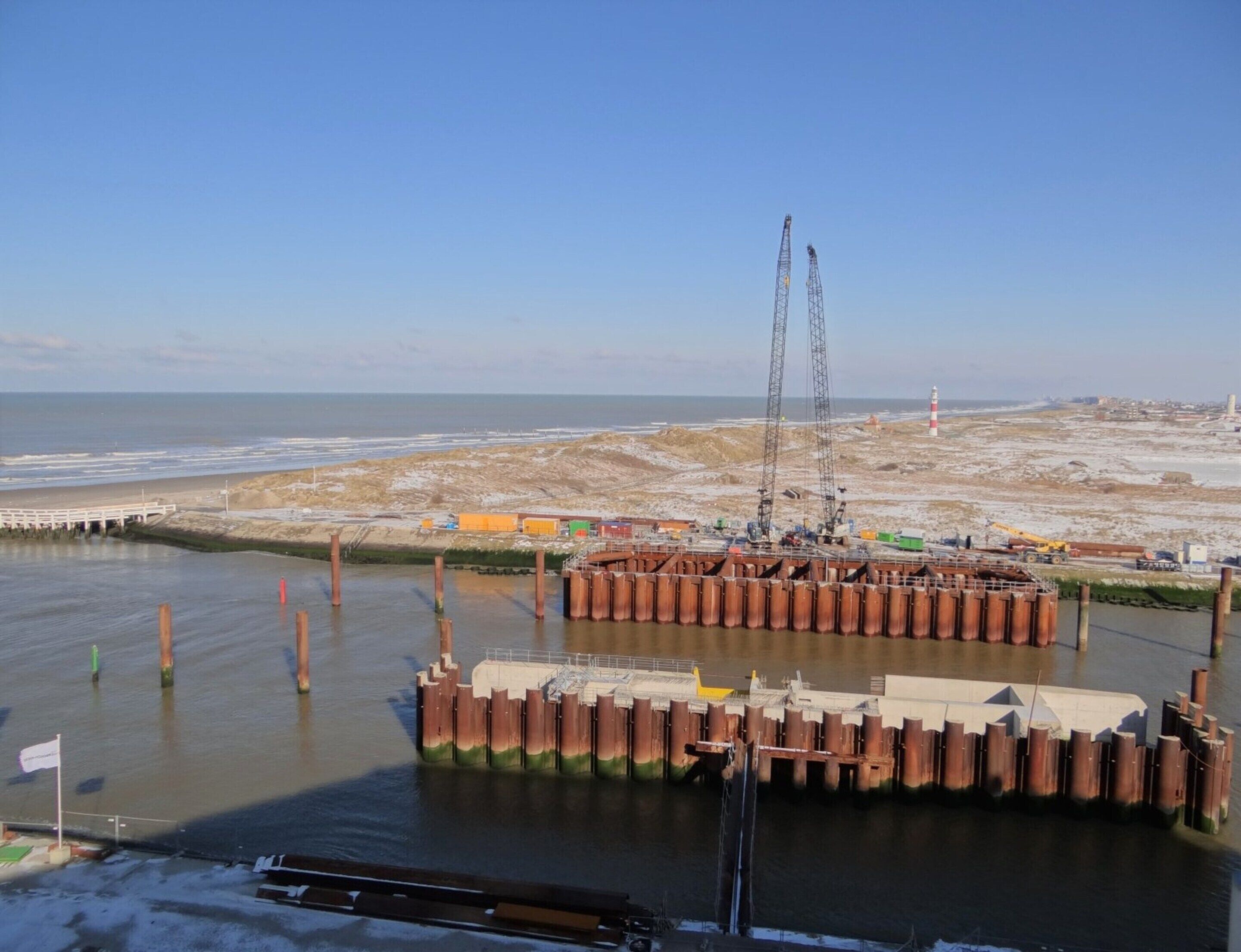 A coastal construction site with a crane, a pier, and a lighthouse in the background.