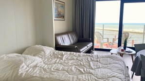A hotel room with a bed, a leather sofa, a small table with a plant, and a view of the sea.