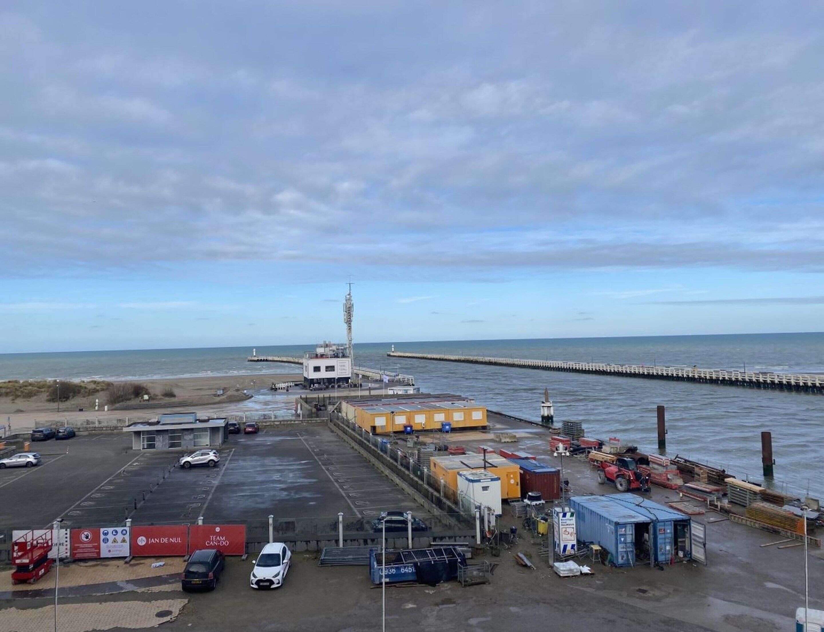 A coastal industrial area with a pier extending into the sea, a small building with a tower, and a parking lot with various vehicles and shipping containers.