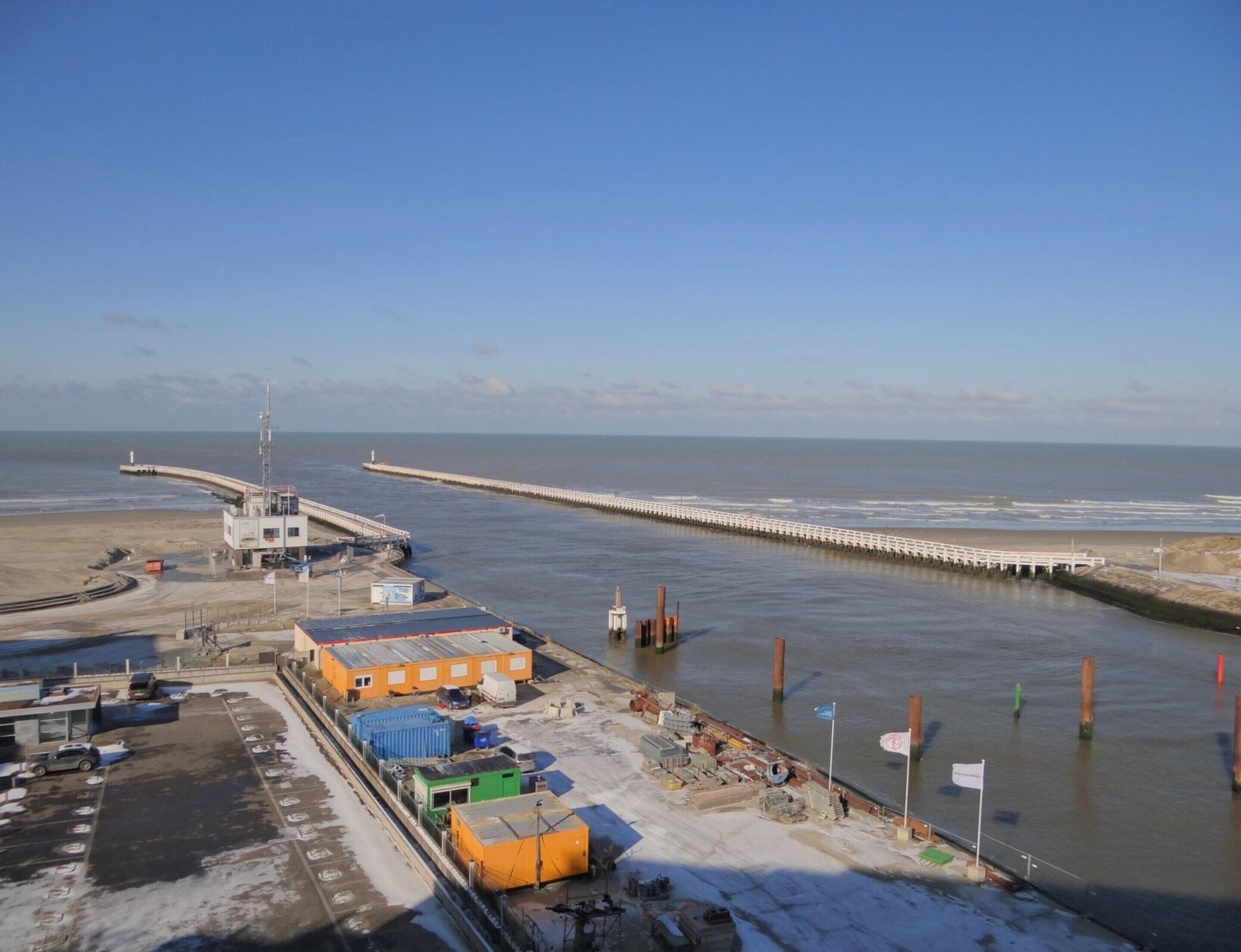 A coastal industrial area with a pier extending into the water, a control tower, and a parking lot with shipping containers.