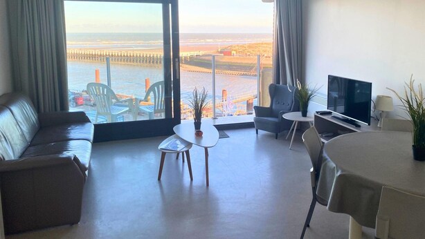 A modern hotel room with a large window offering a view of the sea and a pier, a leather sofa, a small round table with a plant, a TV, and a chair.