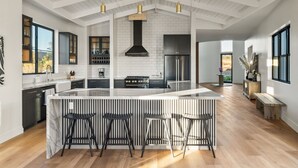 A modern kitchen with a large island, bar stools, and a central chimney. - Vintner's Village | California-Chic Wine Estate (Temecula)