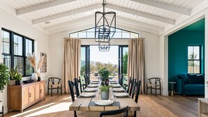 A dining area with a long table set for six, surrounded by chairs, with a view of the outdoors through large windows. - Vintner's Village | California-Chic Wine Estate (Temecula)
