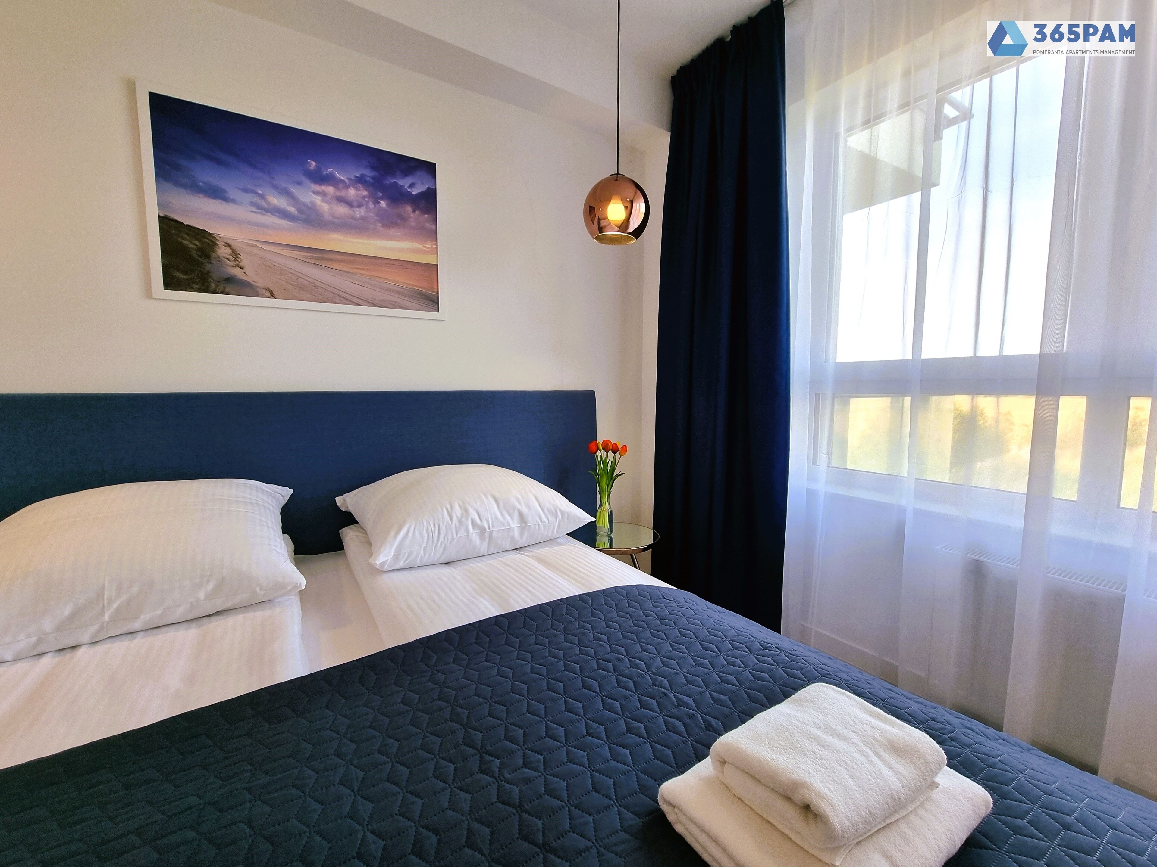 A hotel room with a large bed, two pillows, a nightstand with a vase of flowers, a painting on the wall, and a window with sheer curtains.