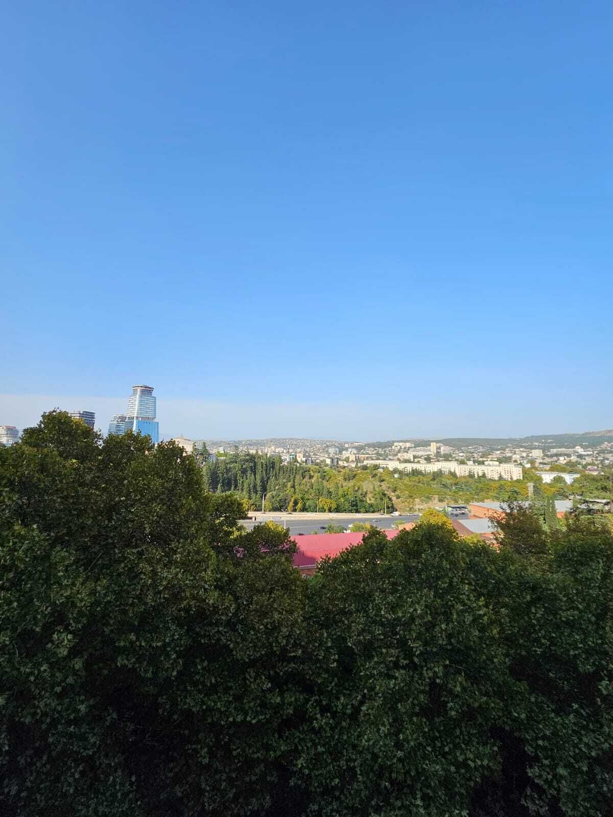 A cityscape with a prominent modern building, dense greenery, and a clear sky.