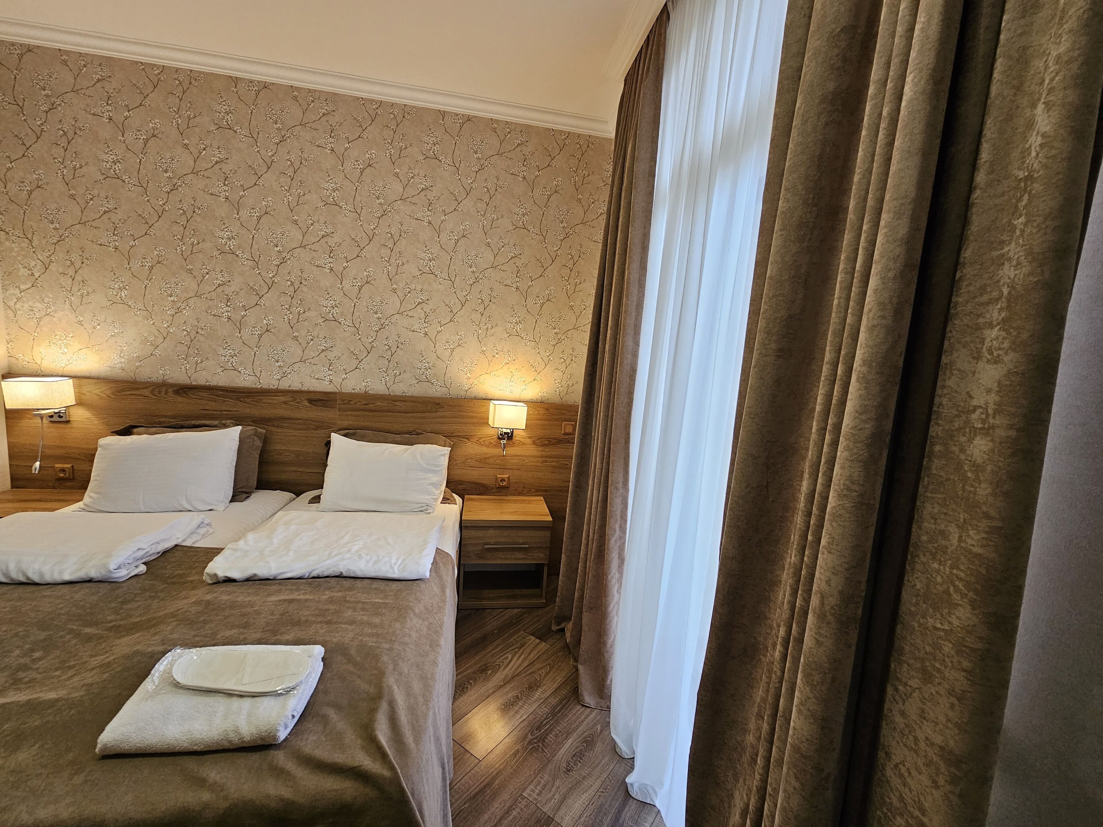 A hotel room with a large bed, two bedside tables with lamps, a wooden headboard, and a window with curtains.