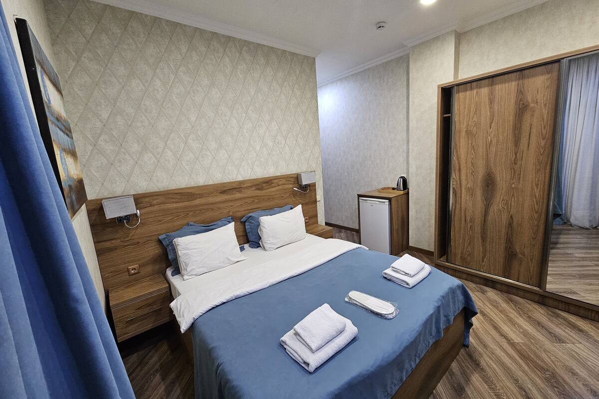 A hotel room with a wooden headboard, a bed with blue bedding, a small bedside table with a lamp, a wooden wardrobe, and a mirror.