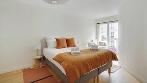 Moderni hotellihuone, jossa on suuri sänky, yöpöytä, lamppu ja suuri ikkuna, josta avautuu näkymä kaupunkiin.