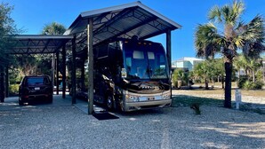 A bus parked under a covered shelter with a gravel ground, palm trees, and a clear blue sky in the background. - RV LOT Pole barn is fully covered and concreted Indian Pass (Port Saint Joe)