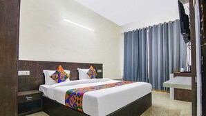 A hotel room with a large bed, a bedside table, a nightstand, a built-in shelf, and a bathroom area with a sink and mirror. - Hotel Viraj (Gurugram)