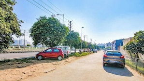 A road with parked cars, trees, and buildings in the background. - Hotel Viraj (Gurugram)