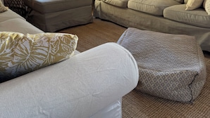 A cozy living room with a beige sofa, a patterned ottoman, and a large potted plant.