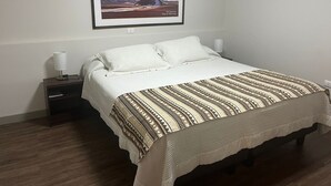 Un lit bien fait, recouvert d’une couvre-lit à motifs, un tableau encadré représentant un paysage de montagne accroché au mur, et une lampe de chevet posée sur une table de nuit.