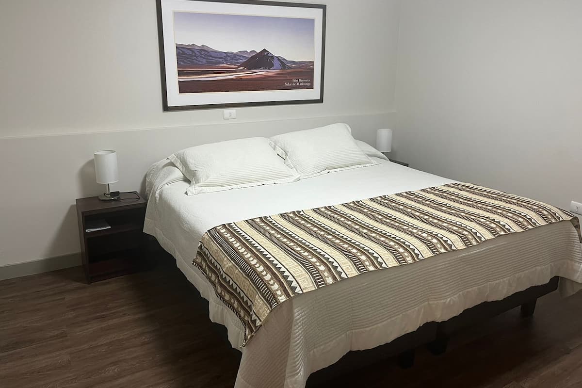A neatly made bed with a patterned bedspread, a framed picture of a mountain landscape on the wall, and a bedside lamp on a nightstand.