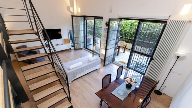 A spacious living area with a wooden staircase, a sofa, a dining table with chairs, and large windows offering a view of the outdoors. - VivimiHome-Esclusivo loft con terrazzo (Milan)