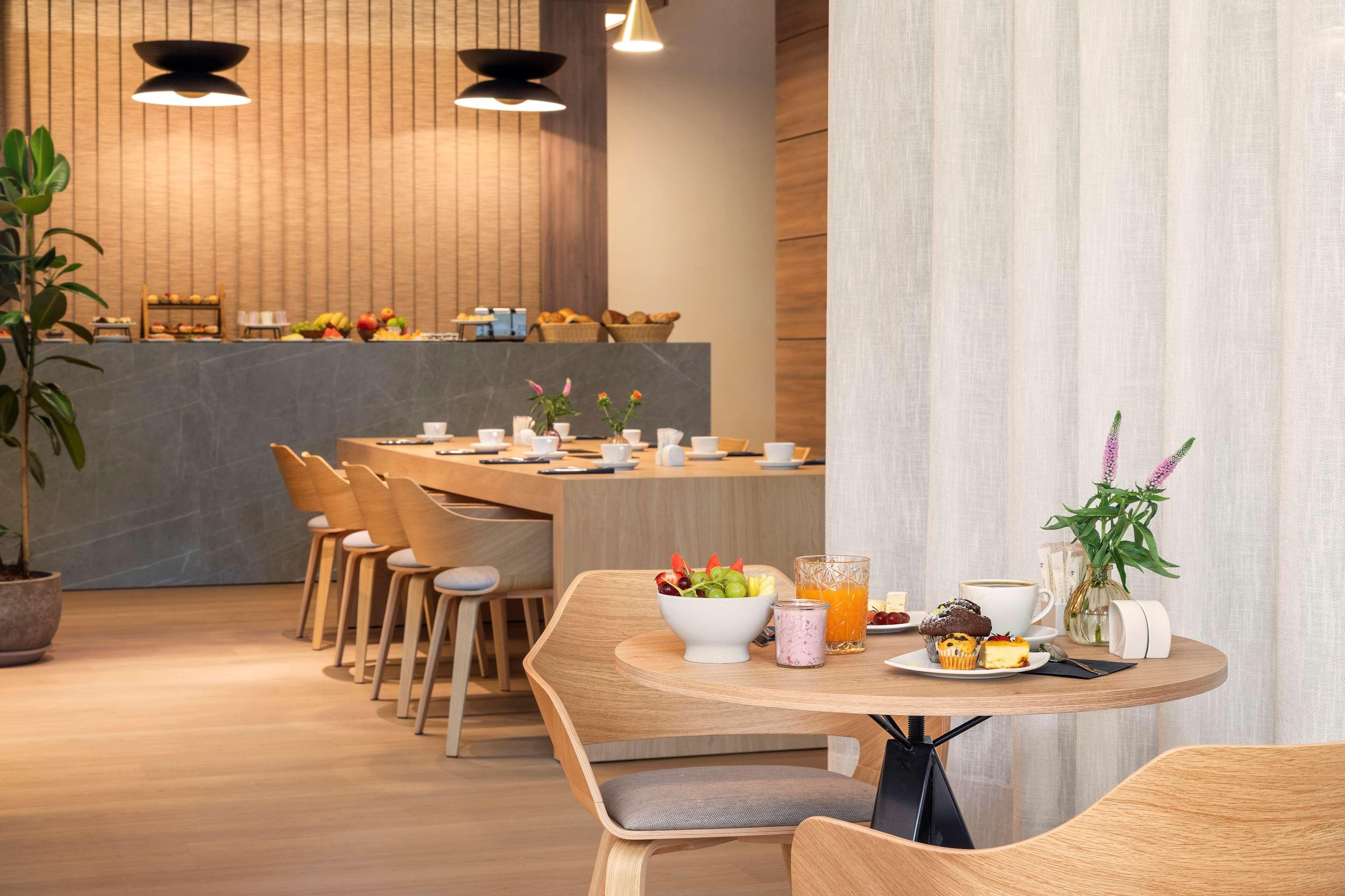A modern dining area with wooden furniture, a central round table with a bowl of fruit and drinks, and a kitchen in the background.