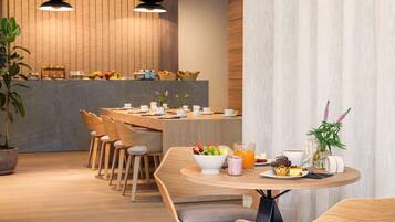 Uma área de refeições moderna com mobiliário de madeira, uma mesa redonda central com uma tigela de frutas e bebidas, e uma cozinha ao fundo.