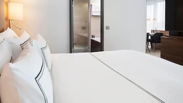 Habitación de hotel con una cama grande, una mesita de noche, una lámpara, zona de ducha y comedor con televisor.