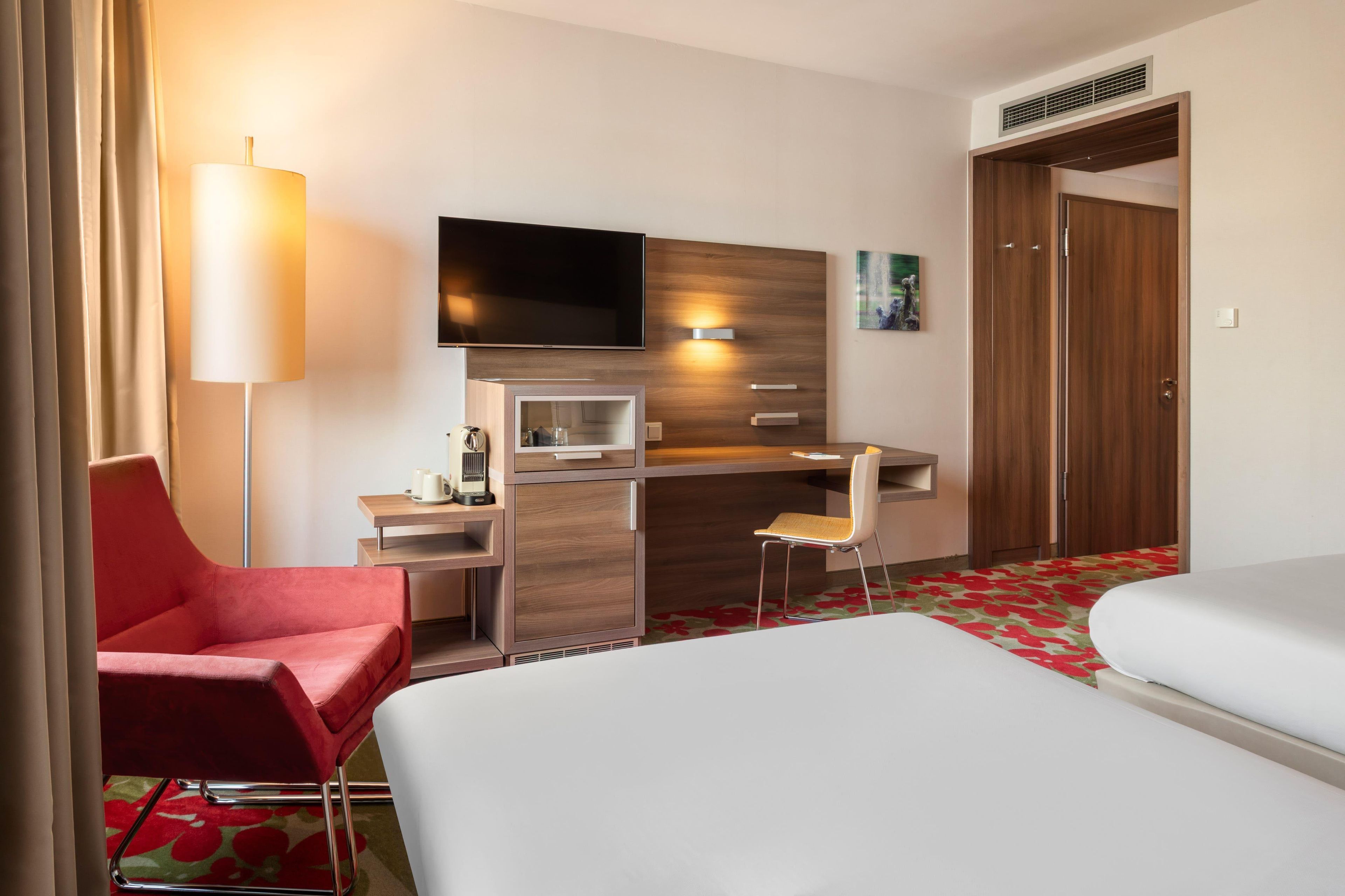 Une chambre d’hôtel comprenant un lit, un bureau avec un téléviseur à écran plat, une chaise rouge, une petite table et une armoire.