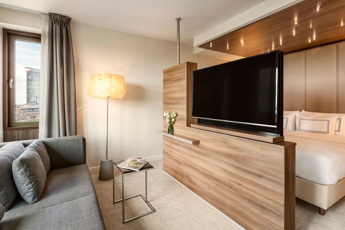 Ένα σύγχρονο δωμάτιο ξενοδοχείου με έναν γκρι καναπέ, μια τηλεόραση επίπεδης οθόνης τοποθετημένη σε έναν ξύλινο τοίχο, ένα φωτιστικό δαπέδου και ένα κρεβάτι με κεφαλάρι.