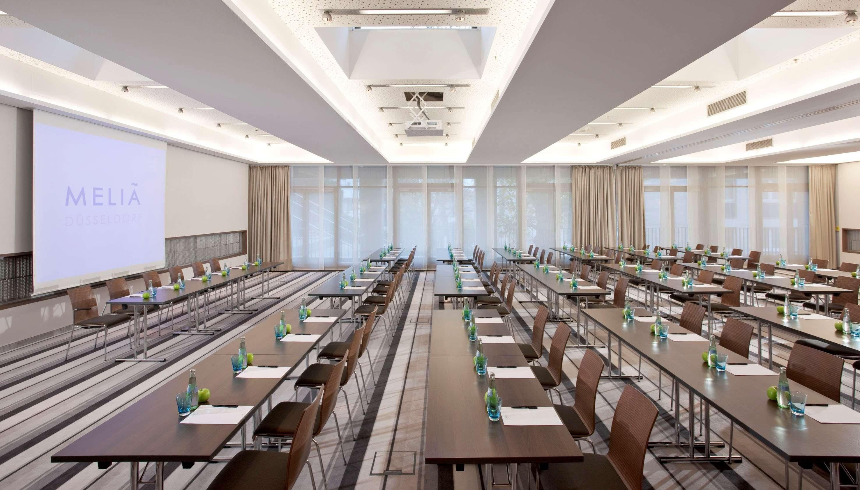 Een congreszaal met lange tafels, stoelen en een groot scherm waarop 'MELIA DÜSSELDORF' staat.