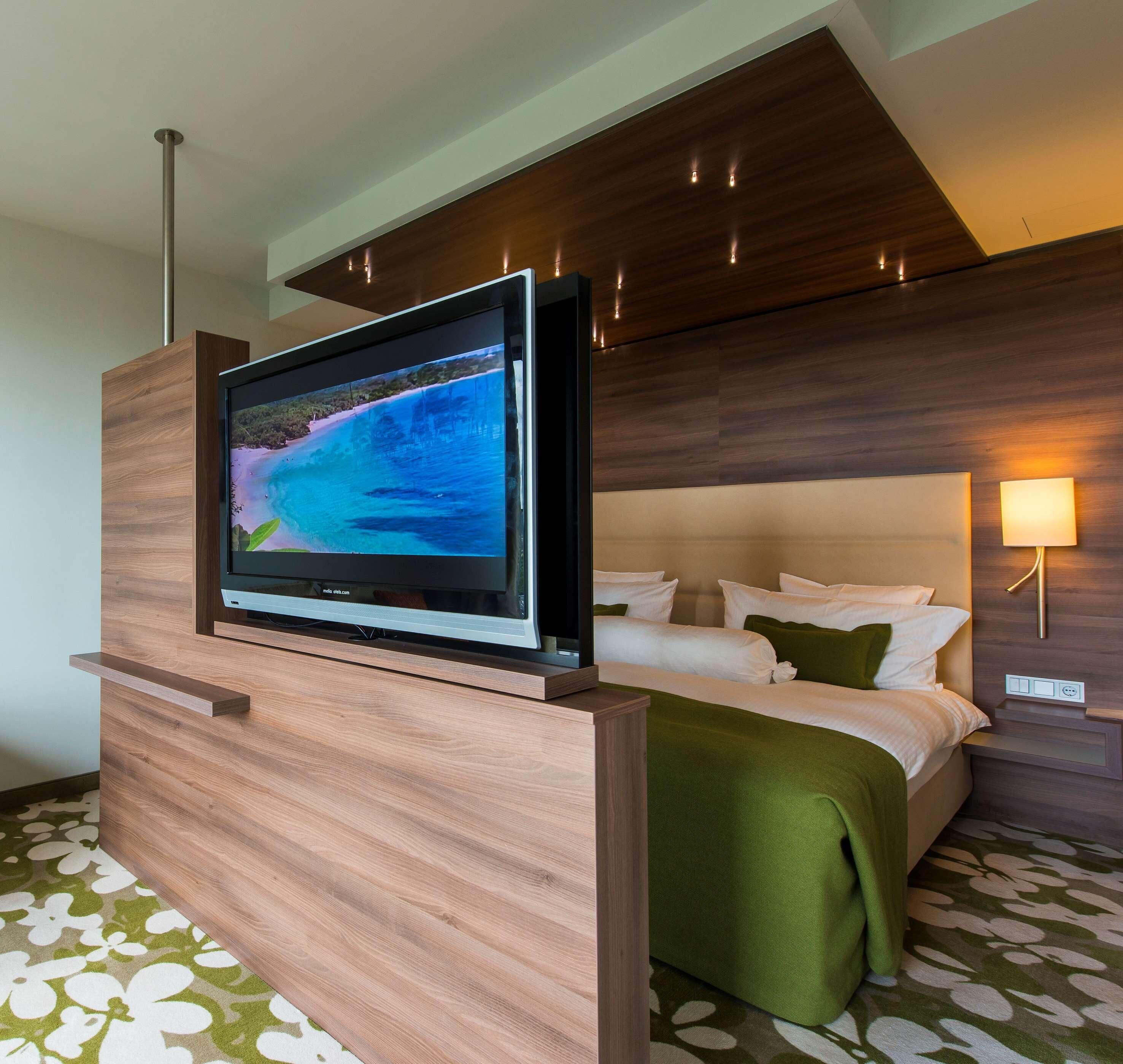 A hotel room with a wooden TV stand, a bed with white and green bedding, and a wall-mounted lamp.