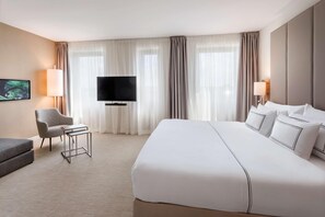 A hotel room with a large bed, a flat-screen TV, a chair, a small table, and a TV on the wall. - Meliá Düsseldorf (Düsseldorf)