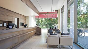 Uma área de recepção moderna com um balcão de madeira, espaço de estar com sofás e janelas amplas com vista para o exterior.