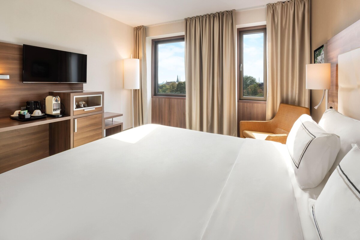 A hotel room with a bed, a desk with a TV, a chair, a window with curtains, and a lamp.