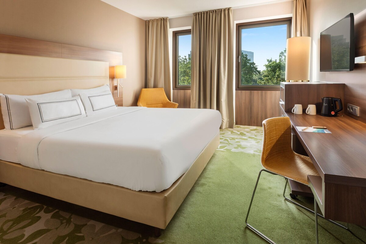 Ein Hotelzimmer mit einem großen Bett, einem Schreibtisch mit Stuhl, einem Fernseher und einem Fenster mit Vorhängen.
