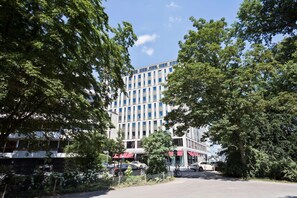 A multi-story building with a modern architectural design, surrounded by lush greenery and parked cars. - Meliá Düsseldorf (Düsseldorf)
