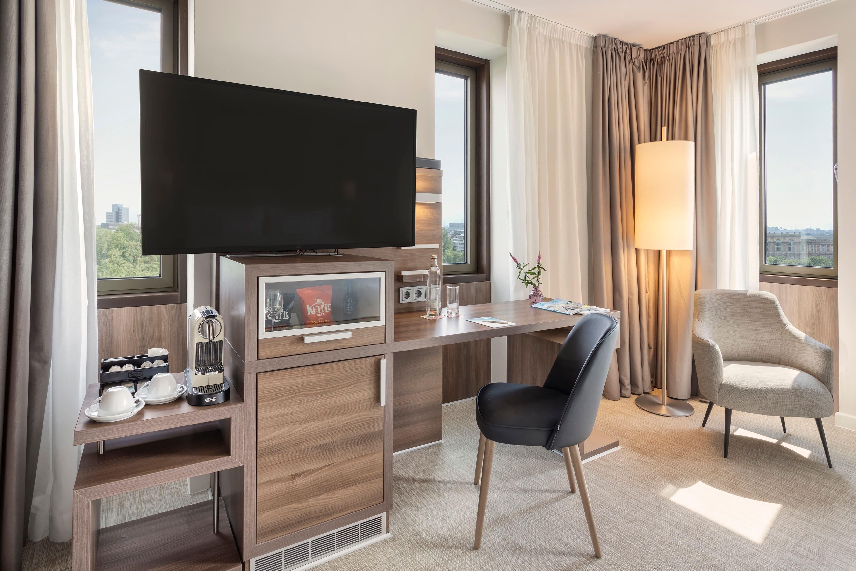 Quarto de hotel com televisão, escrivaninha, duas cadeiras, abajur e vista para a cidade.