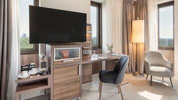 Quarto de hotel com televisão, escrivaninha, duas cadeiras, abajur e vista para a cidade.