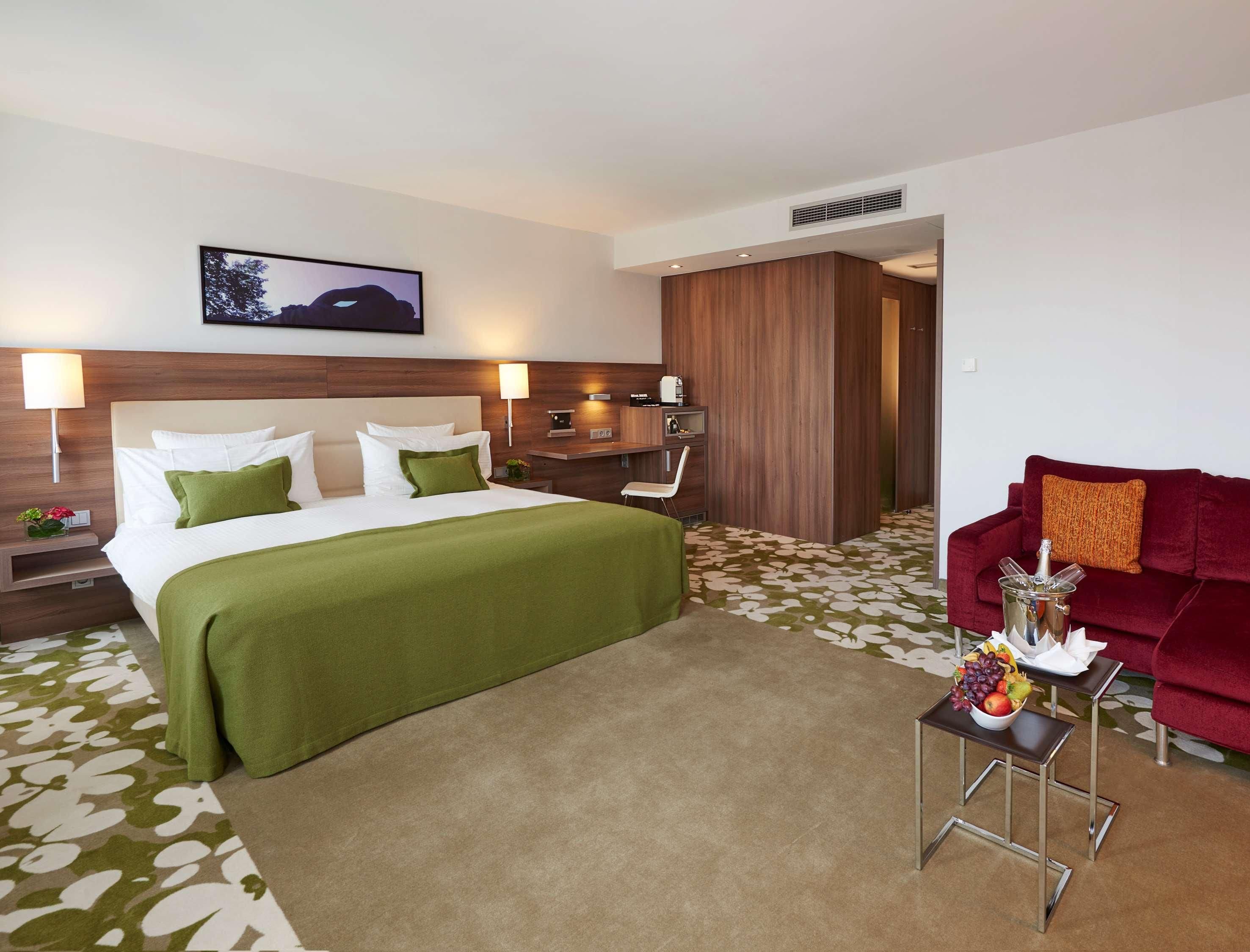Ein Hotelzimmer mit einem Bett, Nachttischen, einem Schreibtisch, einem roten Sofa und einer Obstschale.