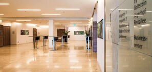 Eine moderne Lobby mit einer Glaswand, die Zimmernamen zeigt, einem Empfangsbereich mit Theke sowie einem sauberen, gefliesten Boden.