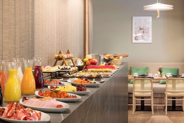 A buffet with various food items and beverages, a sofa, and two chairs in a modern room. - Meliá Düsseldorf (Düsseldorf)