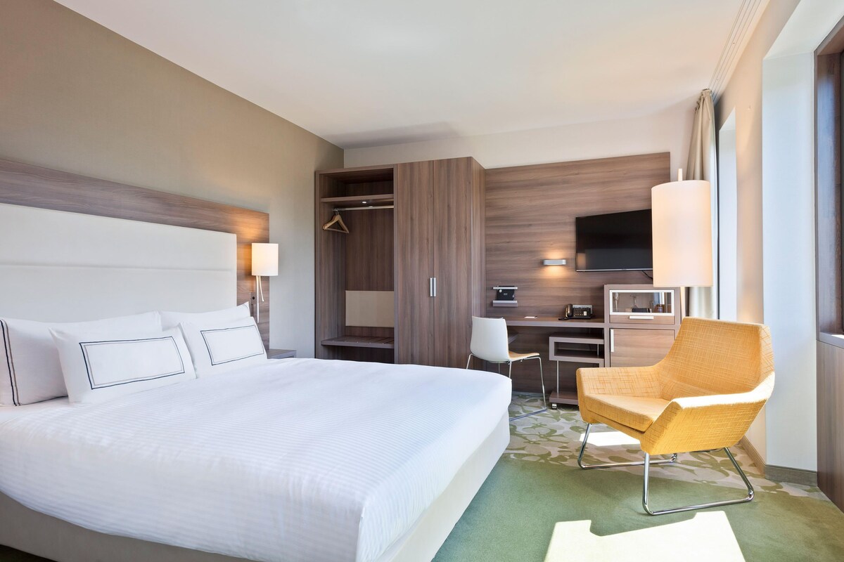 A hotel room with a large bed, a desk, a chair, a TV, and a wardrobe.