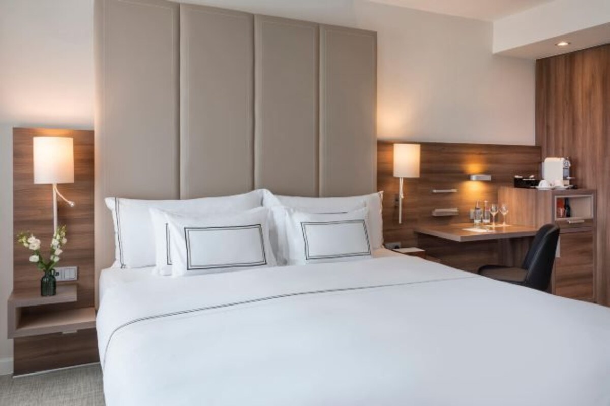 A modern hotel room with a large bed, bedside tables, a desk, and a wooden headboard.