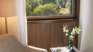Una habitación con vistas a un parque, una silla, una mesita con un florero y una lámpara.