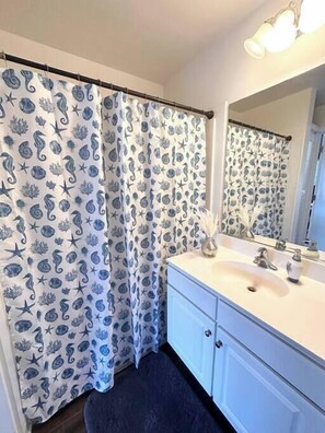 Un baño con cortina de ducha con estampado marino, un lavabo blanco con espejo y una alfombra azul oscuro.