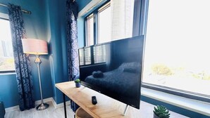 A modern room with a large flat-screen TV, a wooden desk, a chair, a lamp, and a window with curtains. - Your home away from home - Weekly (Charlotte)
