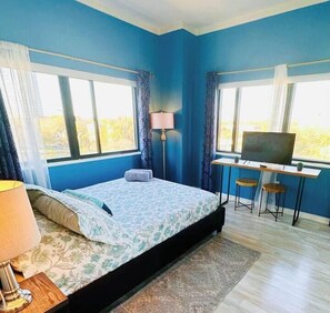 Un dormitorio con cama, escritorio, televisor y ventanas amplias.