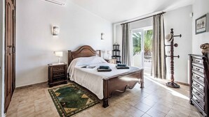 Un dormitorio con una cama de madera, una mesita de noche, una lámpara, un aparador y vistas del exterior a través de una ventana con cortinas.