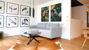 A modern living room with a grey sofa, a round coffee table, and a cowhide rug. There are abstract paintings and a colorful parrot painting on the wall.
